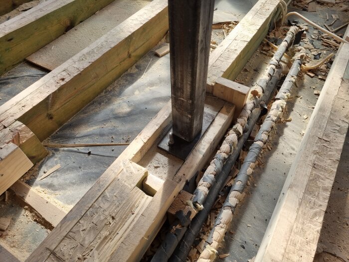 Pelare på gjutet fundament mellan träbjälkar i konstruktion, omgivet av sågspån och några kablar eller rör.