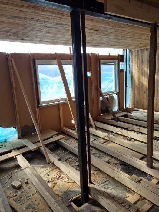 Byggarbetsplats med träreglar, stödbjälkar och två fönster. En pelare står centrerad, omgiven av löst material och träbitar på golvet.