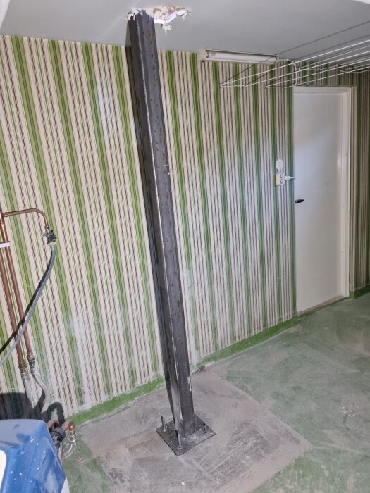 En stålpelare installerad i ett källarutrymme, fastsatt i betonggolvet, med en perforerad grön och vit randig tapet bakom.