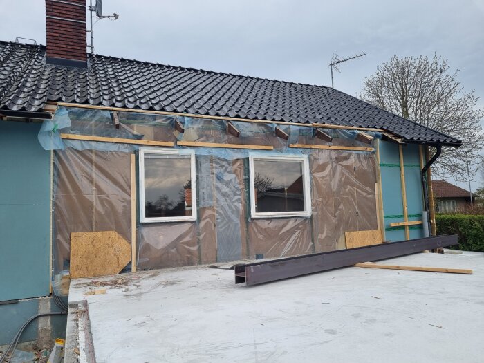 Husfasad med plastskyddade väggar, fönster mitt på väggen, synlig takpanna, skorsten till vänster, balkar placerade på marken framför.