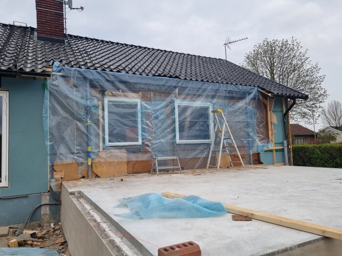 Hus under renovering med plast över fönstren, stegar vid fasaden och en gjuten betongplatta framför. Skorstenen syns på taket.