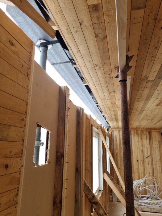 Inre byggkonstruktion med träpaneler, balkar och pelare som stöder en takbalk. En del av en vägg under konstruktion med öppning och fönster.