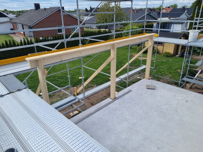 Träkonstruktion under byggnation på husets övervåning, omgiven av byggnadsställningar, med utsikt över en trädgård och omgivande hus.