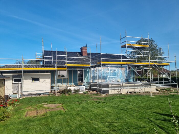 Hus under renovering med byggnadsställningar runt fasaden, i soligt väder. Grön gräsmatta i förgrunden, blå himmel ovanför.