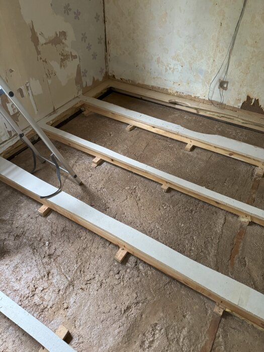 Uppbrutet sovrumsgolv med synlig isolering och reglar, i ett äldre hus byggt 1973. Väggar med bortskavd tapet och sladd längs väggen.