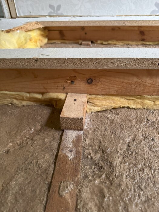 Bilden visar en del av ett golv i ett hus under renovering, med synlig isolering, träreglar och en brun cementplatta med inbäddade träbitar.