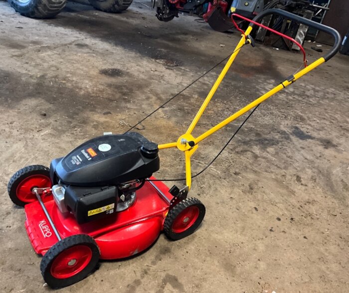 Röd och svart Klippo gräsklippare med gul handtag, stående i ett garage, utan drivning och med en motor från Briggs & Stratton.