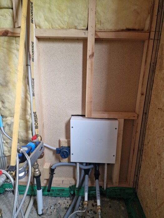 Fördelarskåp monterat i en vägg med exponerade träreglar och isolering, där skåpet sticker ut utanför reglarna. Olika rör och kopplingar visas.