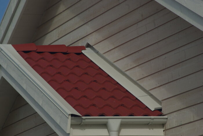Punainen aaltopeltikatto talon kyljessä, jossa on sadevesikouru ja vaalea puupaneeli seinä.