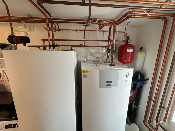 Två bergvärmepumpar med kopparrör och ventiler, en röd trycktank och en behållare på en av pumparna, i en teknisk installation.