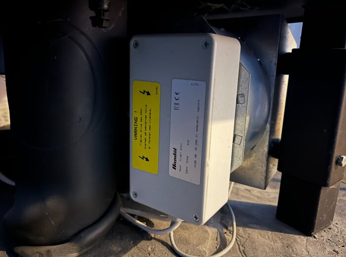 Vit fläktmotor märkt med "Handöl Fläkt 2000" och varningsetikett, monterad under en eldstad. Kablar anslutna under enheten.