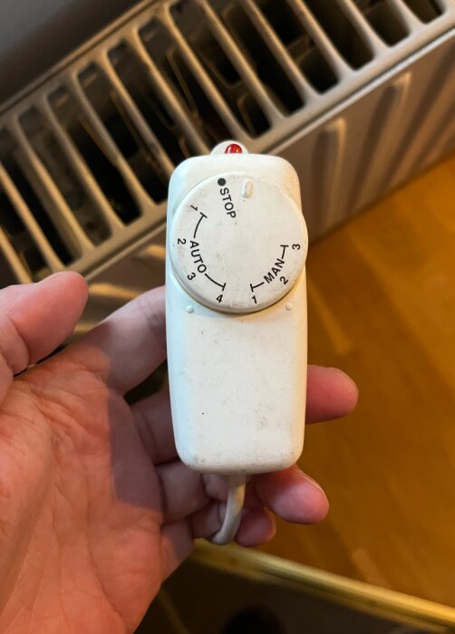 Fjärrkontroll för "Handöl Fläkt 2000" med ratt för auto- och manuella lägen, hålls i en hand bredvid en radiator.