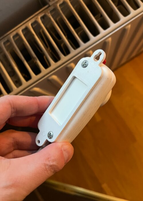 En hand som håller en vit fjärrkontroll nära en radiator. Överdelen av fjärrkontrollen har två skruvar och ett fästhål.