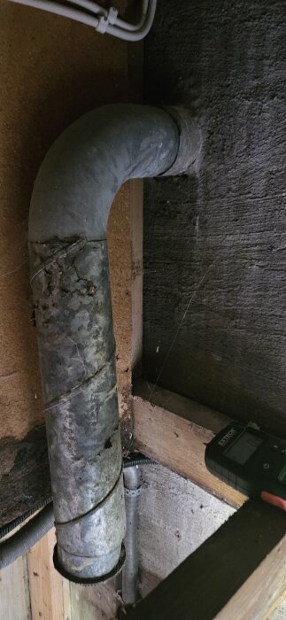 Rostigt metallrör med galler, monterat i hörn av trä- och betongvägg, del av ventilationssystem i äldre radhus.