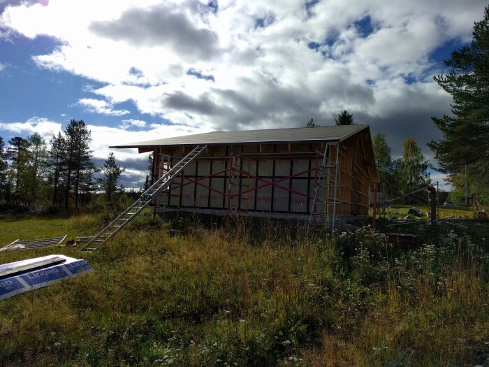 Byggnadsprojekt med ett hus under konstruktion i lantlig miljö, omgiven av träd och gräs, med stegar lutade mot väggarna.