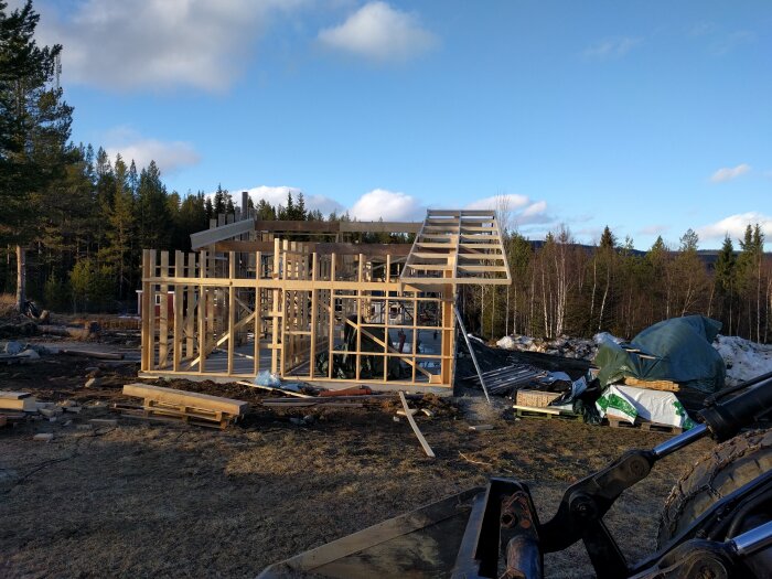 Trästomme till byggnad under uppförande på gräsyta med skog i bakgrunden, diverse byggmaterial och en del av ett fordon synligt.