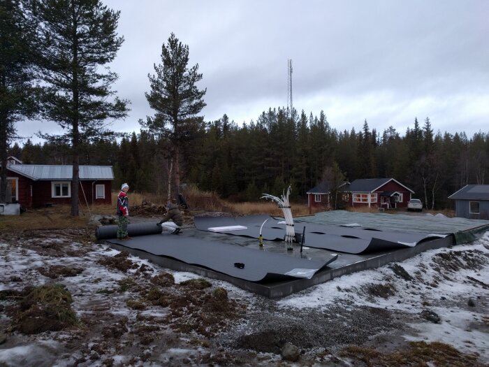 Personer arbetar på en byggarbetsplats med grundläggning i en lantlig miljö, omgivna av träd och trästugor.