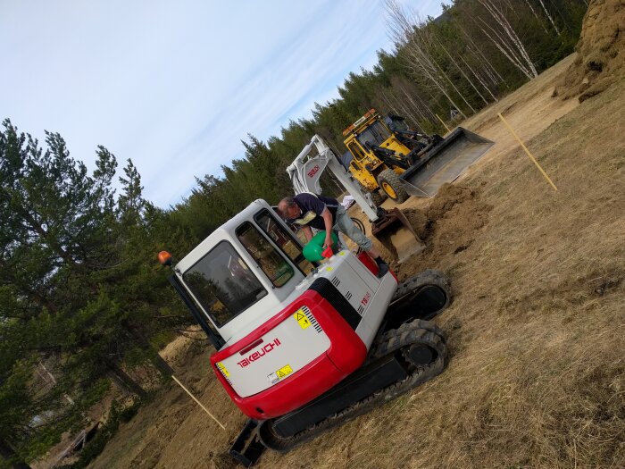 Person fyller på bränsle i en röd och vit grävmaskin på en byggarbetsplats med skog i bakgrunden. En gul lastmaskin syns i närheten.