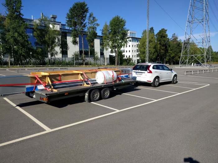Vit SUV parkerad på asfalt med släpvagn som transporterar byggmaterial och fat, bakgrund med kontorsbyggnader och träd.
