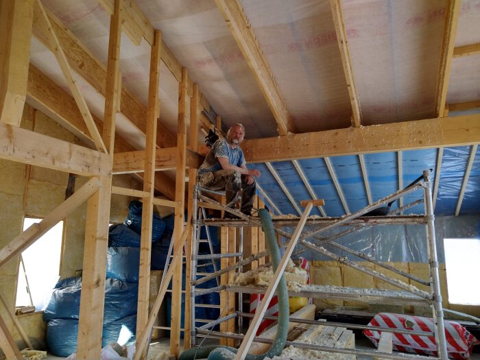 Person som sitter på byggnadsställning i ett hus under konstruktion, med synlig trästomme och isolering.