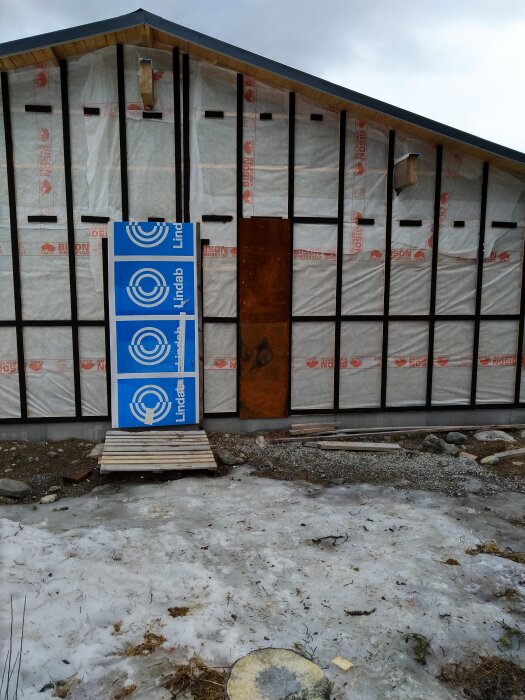 Ett halvt färdigt byggnadsprojekt med väggar täckta av vit isolering och Lindab-paneler, omgiven av snötäckt mark och byggmaterial.