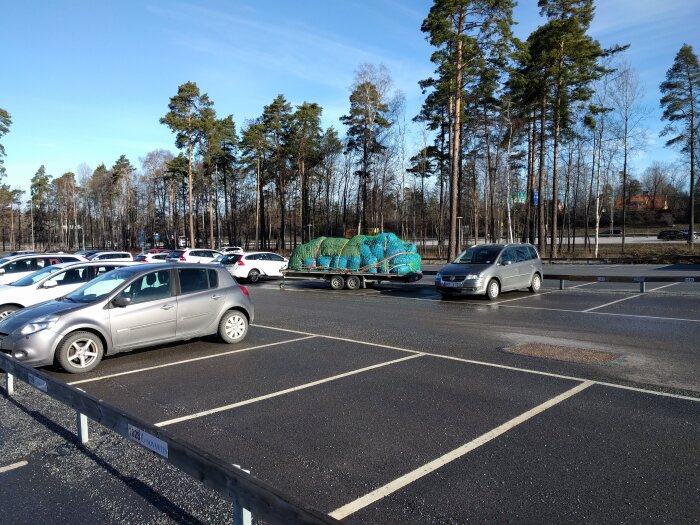 Bilar parkerade på asfaltparkering, en bil fraktar en släpvagn lastad med inplastade säckar, omgivna av höga tallar under blå himmel.