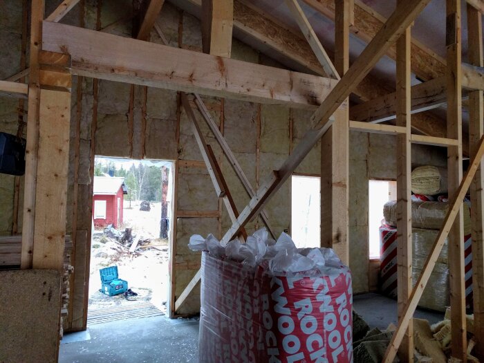 Byggarbetsplats med synliga träreglar, isolering och öppning mot utsidan där en röd stuga syns. Byggmaterial och verktyg på golvet.