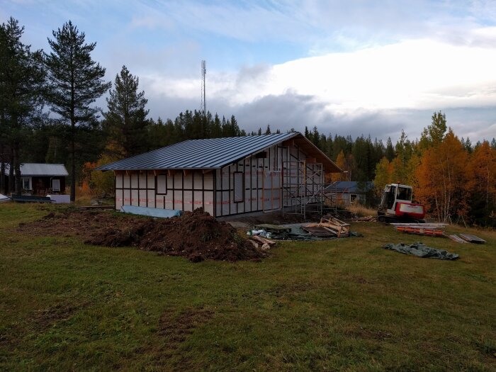 Byggnad under konstruktion med stålramstak och isolerade väggar på en grön tomt, omgiven av skog och en grävmaskin i närheten.