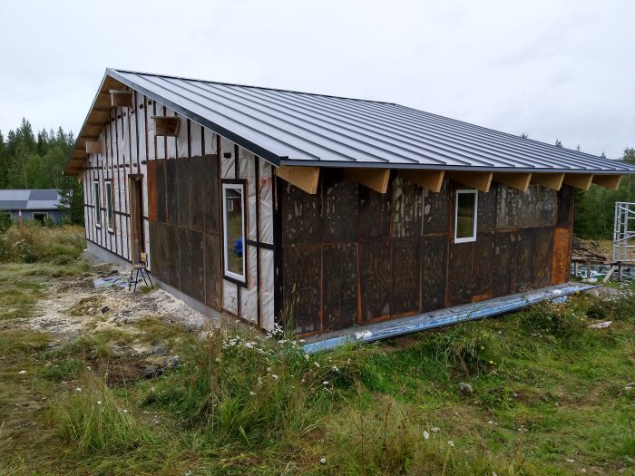 Ett halvfärdigt hus under konstruktion på en grönskande tomt, med synlig isolering och oklara väggar samt grått plåttak.