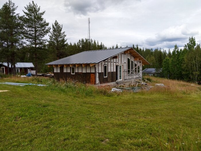 Ett halvfärdigt hus med trä- och metalltak står på en grön gräsplan omgiven av träd. Byggställning synlig vid ingången.