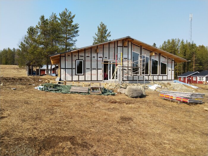 Trähus under byggnation med monterade fönster, ställning runt huset och material utspritt på marken, omgiven av skog och gräsmark.