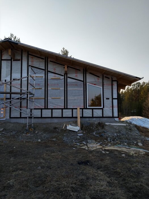 Hus under konstruktion med synlig byggställning, isoleringsmaterial och delvis uppsatt fasad, omgiven av natur och markarbete.