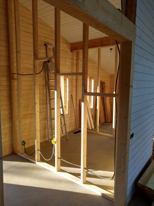 Inre av en pågående husbyggnation med träväggar, synliga reglar och en stege mot väggen. Solens ljus skapar skuggor på golvet.