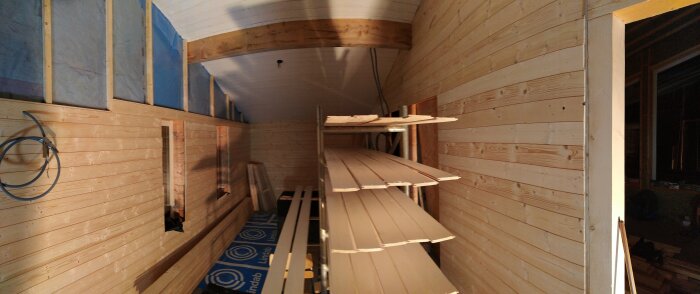 Träpaneler uppsatta på väggar och tak i ett rum under konstruktion. Träplankor förvarade på ställning längs väggen.