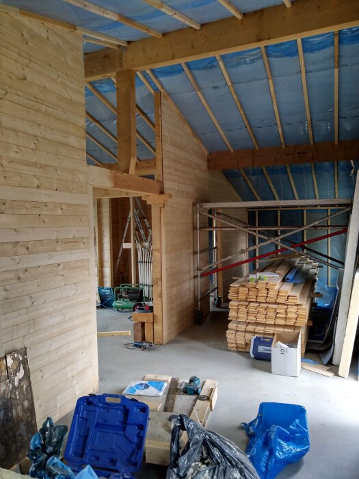 Pågående byggprojekt med träpaneler, verktyg och material staplade inomhus, synliga takbjälkar och blå isolering.
