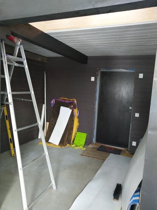 Stege lutad mot väggen i ett rum under renovering med svart dörr, tak med synliga balkar, och byggmaterial samt verktyg på golvet.