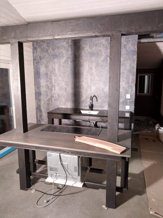 Kök under renovering med bänkskivor i mörkt trä, en diskbänk och en inbyggd spis. Under bänken syns en mikrovågsugn som inte är installerad.