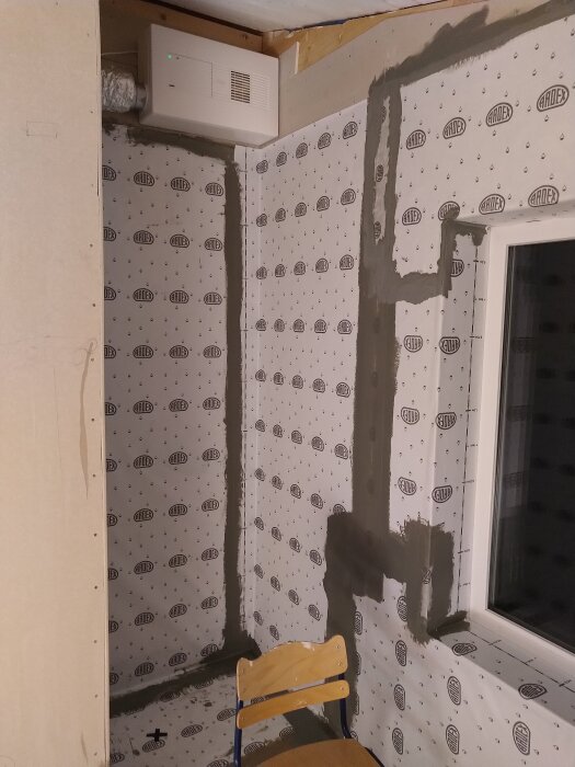 En byggarbetsplats med fuktspärr och kakelväggar på väg att sättas upp, med en stol och en ventilationsenhet i hörnet.
