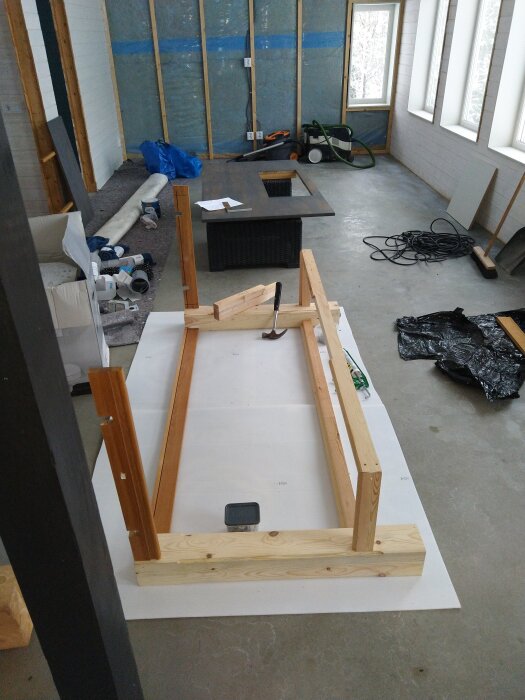 Träram under byggnation på ett golv, omgiven av verktyg, sladdar och byggmaterial i ett rum med isolerade väggar och stora fönster.