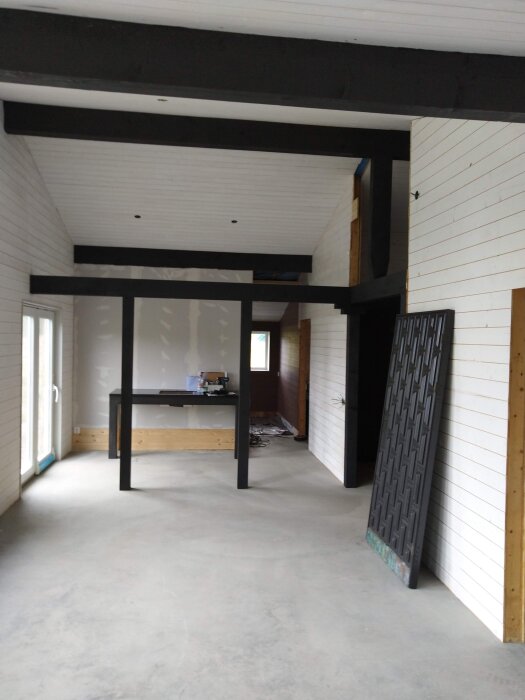 Rymligt rum under renovering med vita träpaneler, svarta bjälkar i taket och betonggolv. Ett arbetsbord och verktyg syns i bakgrunden.