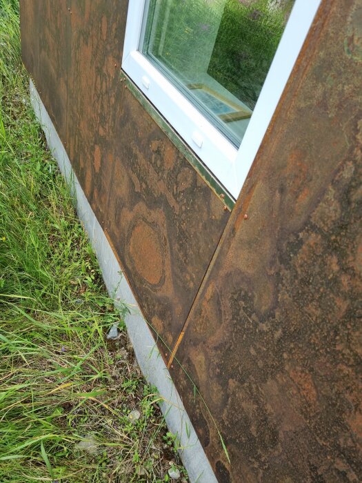 Rostig metallpanel på vägg med vitt fönsterkarm och grön växtlighet vid betonggrund.