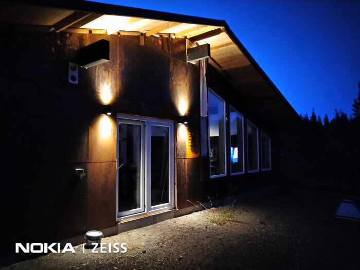 Upplyst husfasad i mörker med stora fönster och trädetaljer.