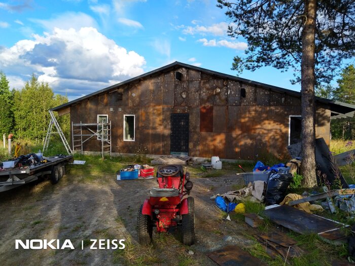 Byggarbetsplats med en halvfärdig fasad av plåtskivor, byggställning, verktyg på släp och omgivande träd.