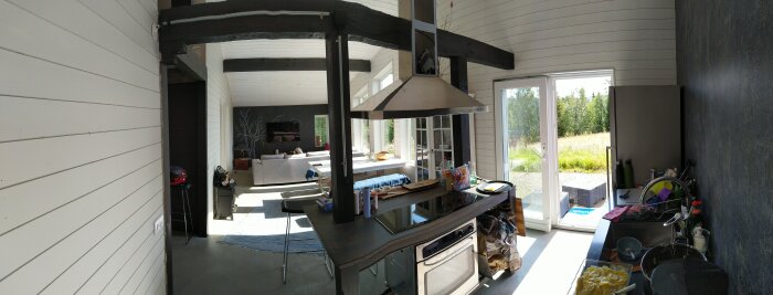 Modernt kök med matplats, stora fönster och utsikt mot trädgård, synlig köksö med spis och ugn, ljus interiör och trägolv.