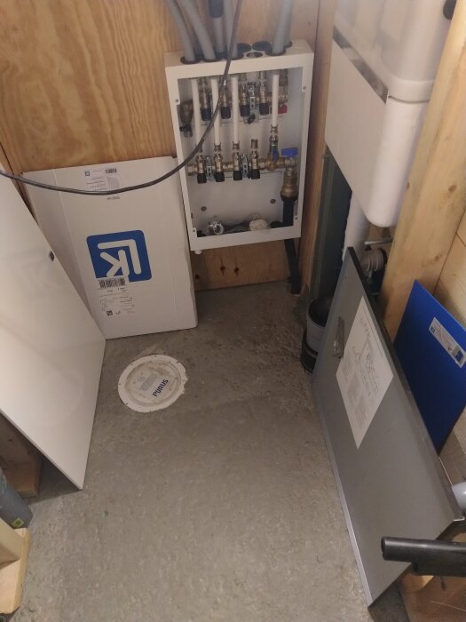 Mekaniskt rum med rörkopplingar på väggen, omgivna av plywoodväggar, ett lock märkt "Purus" på golvet och olika byggmaterial runtom.