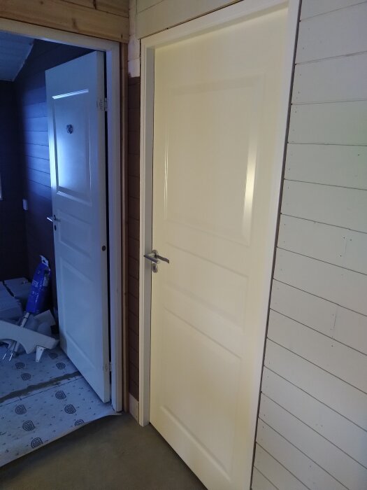 Två vita dörrar i en renoverad interiör, varav en är stängd och den andra är halvöppen mot ett rum med byggmaterial på golvet.