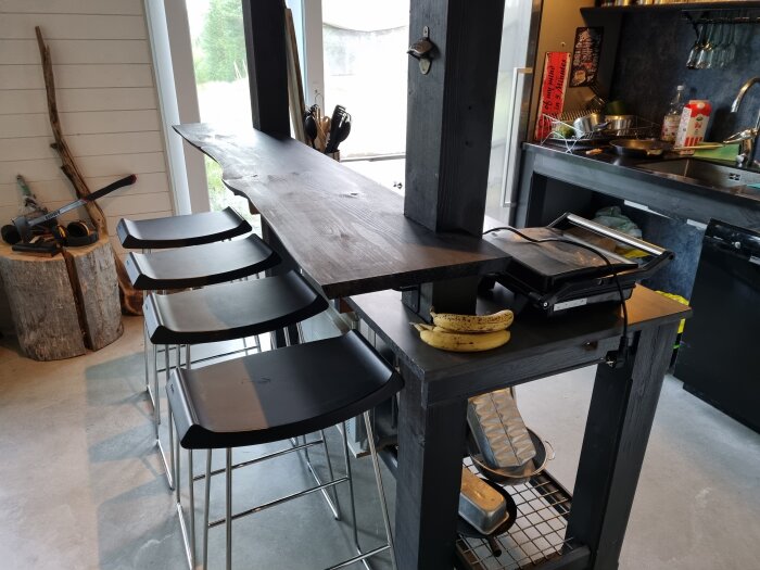 Kök med träbar, tre svarta barstolar, smörgåsgrill, bananer och diverse köksredskap. Rustik stil med trästubbe och ved i bakgrunden.