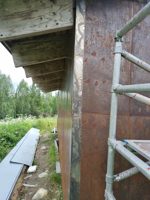 Byggnadsvägg med rostiga plåtskivor, ställning och träbjälkar vid taket. Plåtar på marken och grönska i bakgrunden.