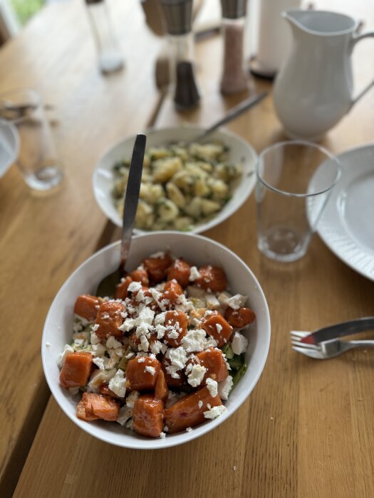 Skål med sallad innehållande lax, avokado, pinjenötter, fetaost och örter. I bakgrunden skål med fransk potatissallad.