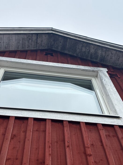 Röd träfasad med ett vitt fönster. Vindskenor och takfot syns högst upp. Färgen har flagnat en del.